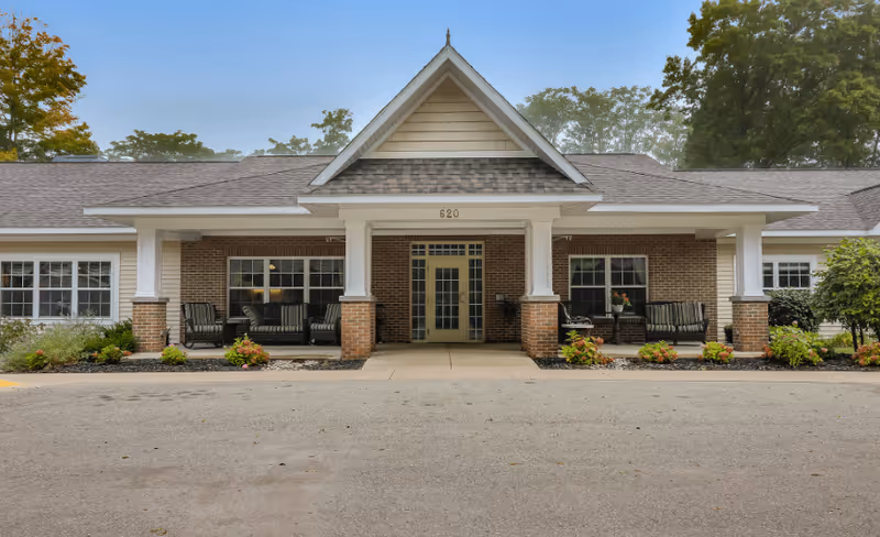 Front exterior view of a single-story brick building with a covered entrance supported by white columns. The entrance has double glass doors and the number 620 above it. There are seating areas with chairs on both sides of the entrance, surrounded by small landscaped plants and shrubs. Trees are visible in the background under a clear sky.