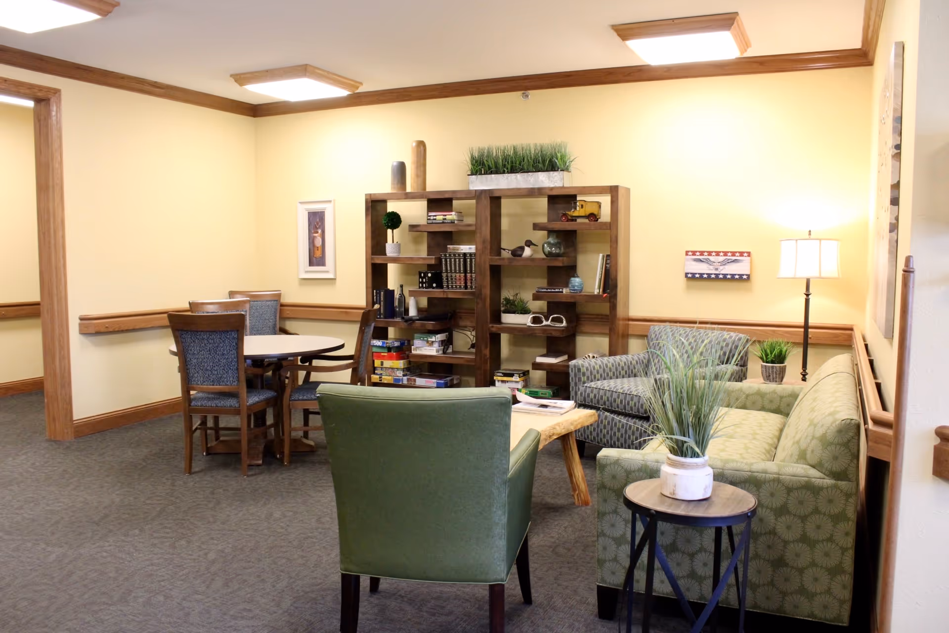 A cozy common area in a senior living facility featuring a green armchair, a patterned green sofa, a small round side table with a potted plant, a wooden coffee table, and a wooden bookshelf filled with books and decorative items. There is also a round table with four chairs in the background, yellow walls with wood trim, a floor lamp, and framed artwork on the walls.