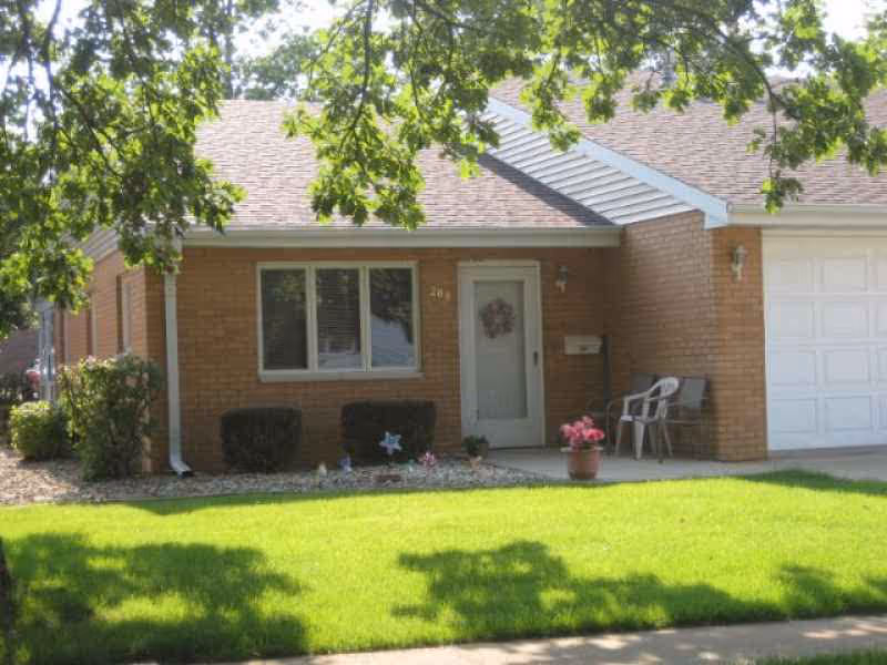 Exterior view of a single-story brick house with a front door, a window, a garage door, and a small porch area with two chairs and a potted plant. The lawn is green and well-maintained with trees providing shade.