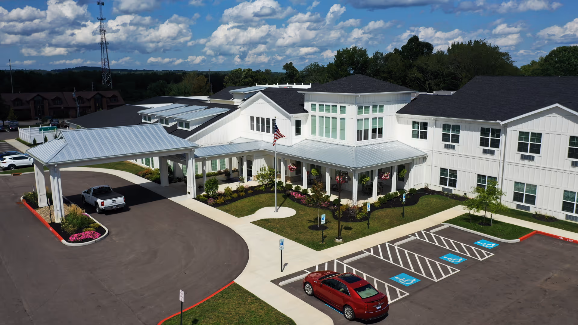 Aerial view of Heritage Crossing senior living facility showing a large white building with a covered entrance, landscaped garden with flowers and an American flag, and a parking lot with several handicap parking spaces and a few cars parked.