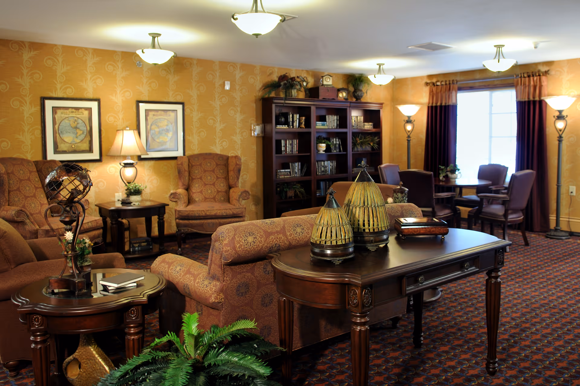 Cozy communal living room with upholstered chairs, wooden tables, a bookshelf, and warm lighting.