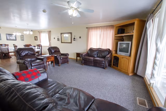 A spacious living room with three dark brown leather sofas and two matching armchairs arranged around a wooden side table. The room has beige walls with framed pictures and decorative stars, carpeted flooring, and large windows with light curtains allowing natural light. A wooden entertainment center with a TV is positioned against one wall. In the background, a dining area with wooden tables and chairs is visible under hanging light fixtures.