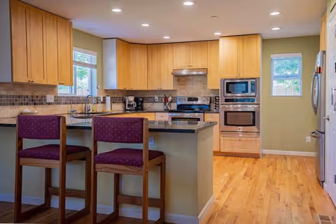 A modern kitchen with wooden cabinets, a granite countertop island with two wooden bar stools with purple cushions, stainless steel appliances including a stove, microwave, and refrigerator, and hardwood flooring. There are two windows letting in natural light.