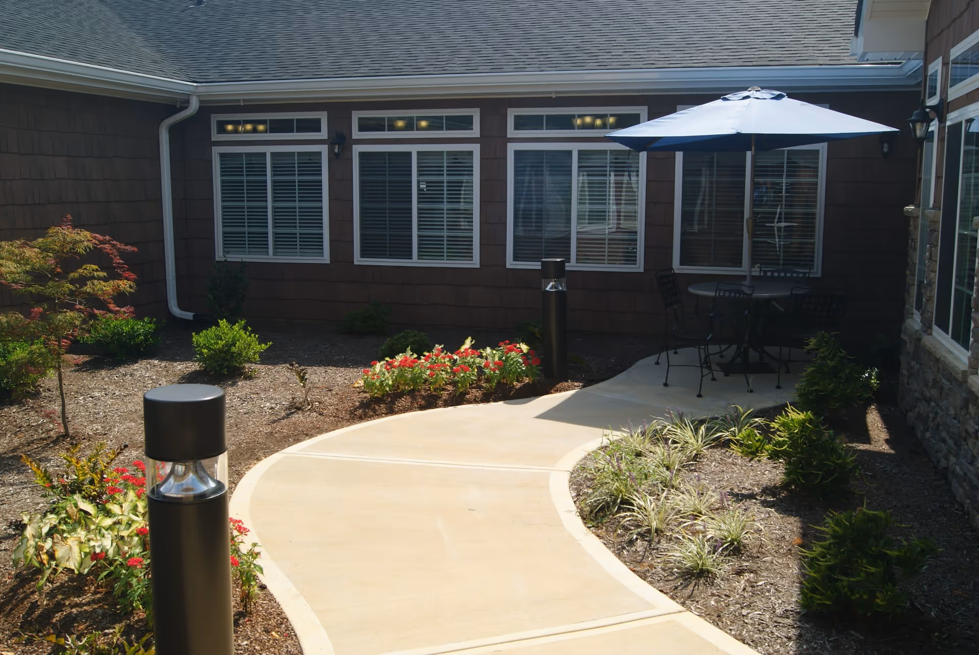 Curved concrete pathway leading to a patio area with a round table and four chairs under a blue umbrella, surrounded by landscaped garden beds with small shrubs and flowers, adjacent to a building with brown siding and multiple windows with white shutters.