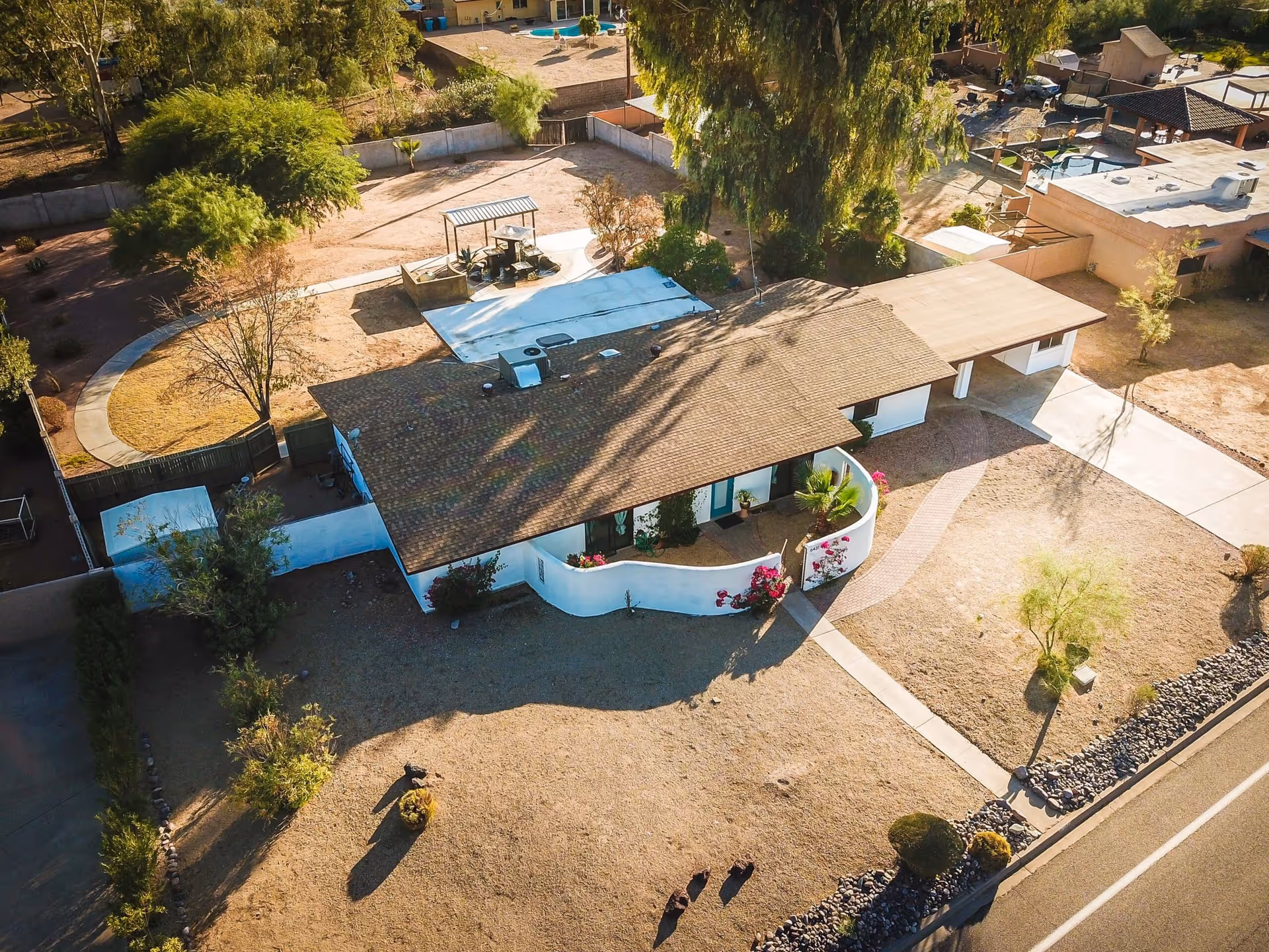 Aerial view of a single-story residential building with a brown shingle roof and white exterior walls, surrounded by a dry landscape with sparse trees and bushes. The property includes a curved walkway, a covered carport, and a fenced backyard area with outdoor seating and a small pool. The building is located next to a road with a sidewalk and rock border.