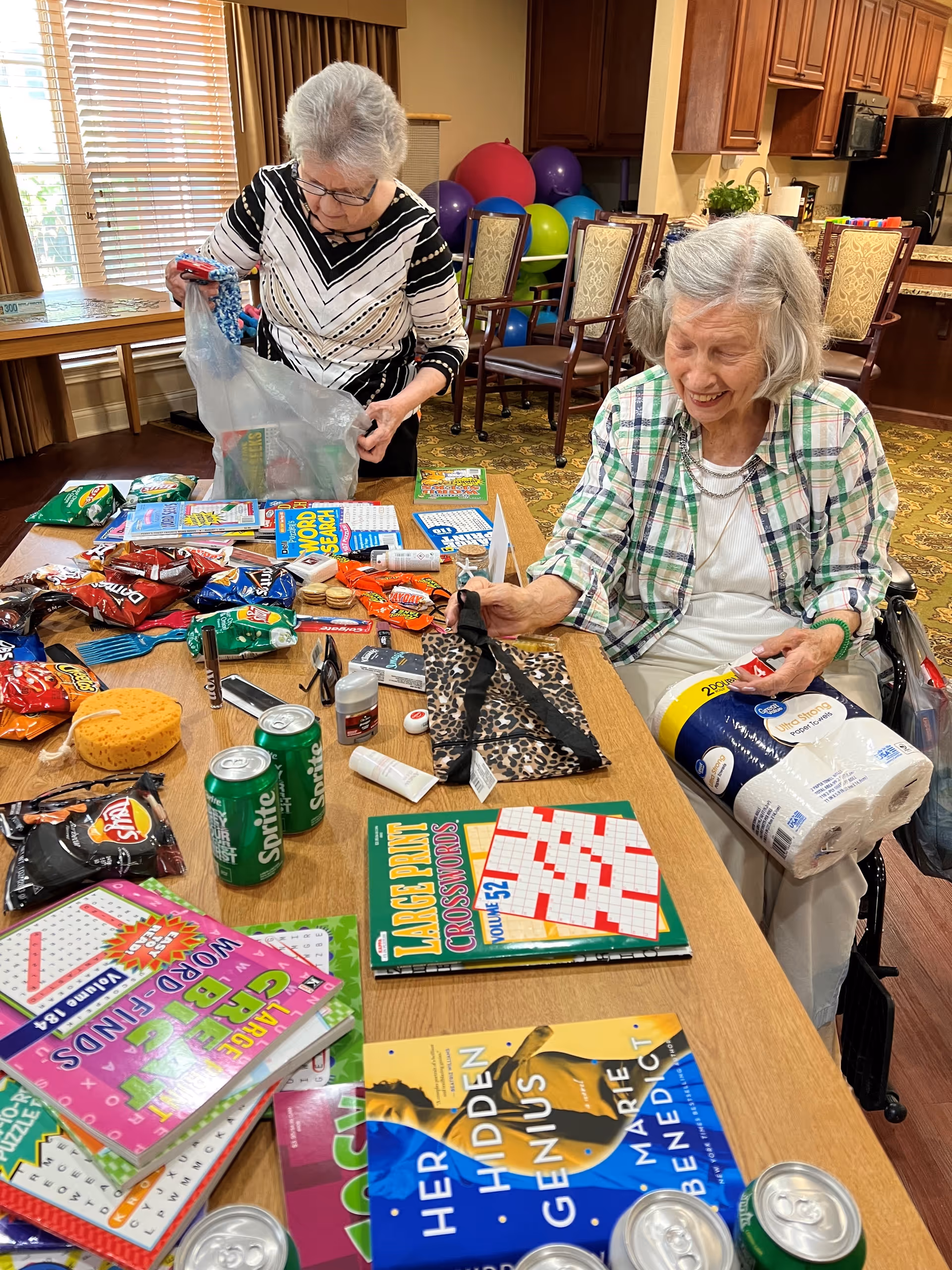 Two elderly women are at a table filled with various snacks, crossword puzzle books, and canned drinks. One woman is standing and holding a plastic bag, while the other woman, seated in a wheelchair, is smiling and holding a package of paper towels. The room has wooden furniture, exercise balls in the background, and a kitchen area visible behind them.