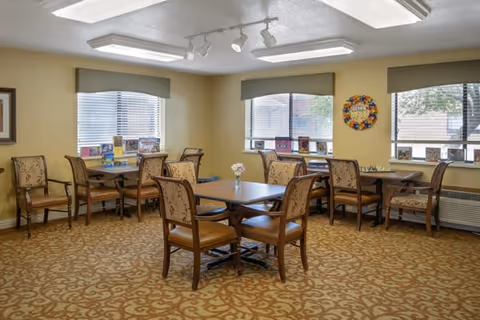 A bright communal dining room with several wooden tables and upholstered chairs arranged near large windows and decorated walls.