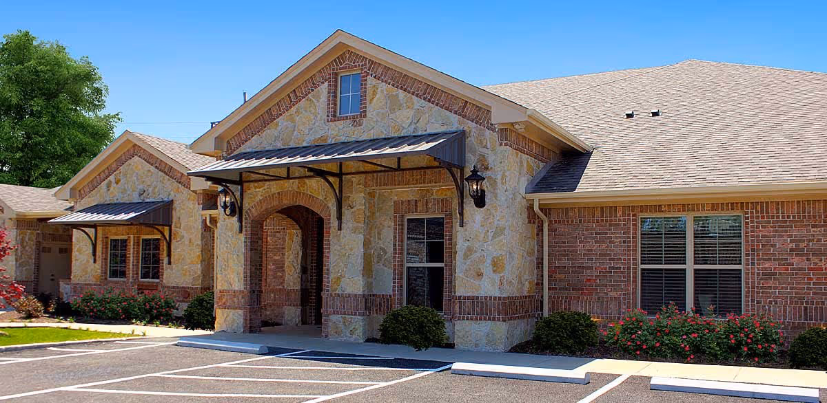 Exterior view of a single-story building with a stone and brick facade, featuring a covered entrance with metal awnings and outdoor wall lanterns. There are windows with blinds and landscaping with bushes and flowers in front, along with a parking area in the foreground under a clear blue sky.