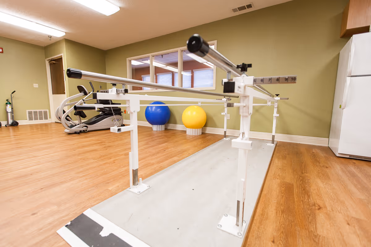 A rehabilitation room with parallel bars for walking exercises, two large exercise balls (one blue and one yellow), an elliptical machine, and a white refrigerator against a green wall. The floor is wooden, and there is a window looking into another room.