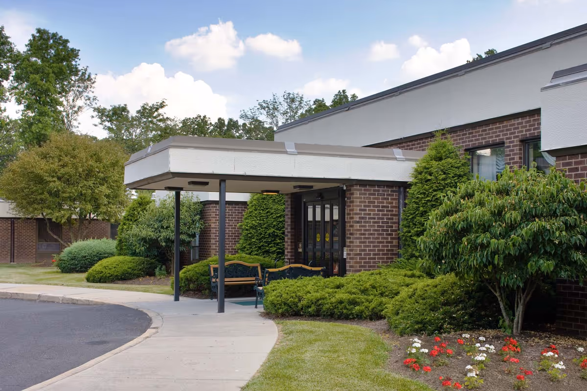 Front entrance of Quakertown Center with a covered porte-cochere, double glass doors, benches, and landscaped shrubs and flowers.