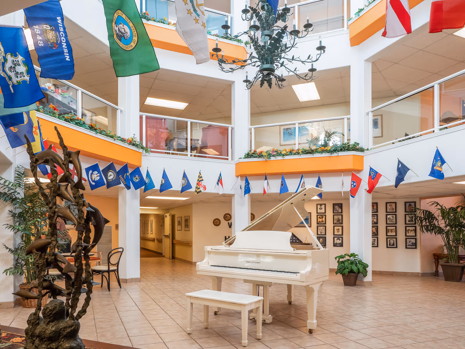 Spacious and bright atrium area inside Holiday Kalama Heights featuring a white grand piano with a matching bench in the center. The space is decorated with numerous state flags hanging from the upper level balcony. There is a large decorative chandelier hanging from the ceiling, plants placed around the room, and framed photos on the walls. The floor is tiled and the upper level has glass railings with orange trim.
