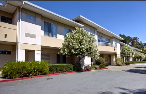 Exterior view of a two-story beige building with several windows and white flowering trees along the sidewalk under a clear blue sky.