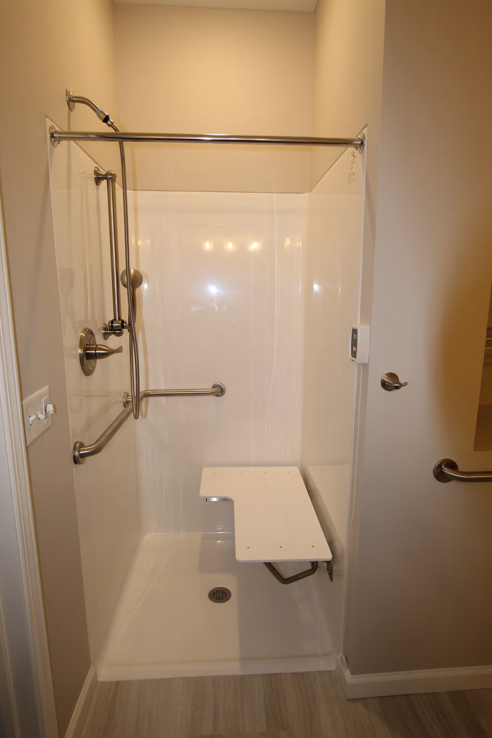 A walk-in shower with a fold-down white seat, stainless steel grab bars, a handheld showerhead, and a fixed showerhead. The shower has a beige wall and light-colored flooring outside.