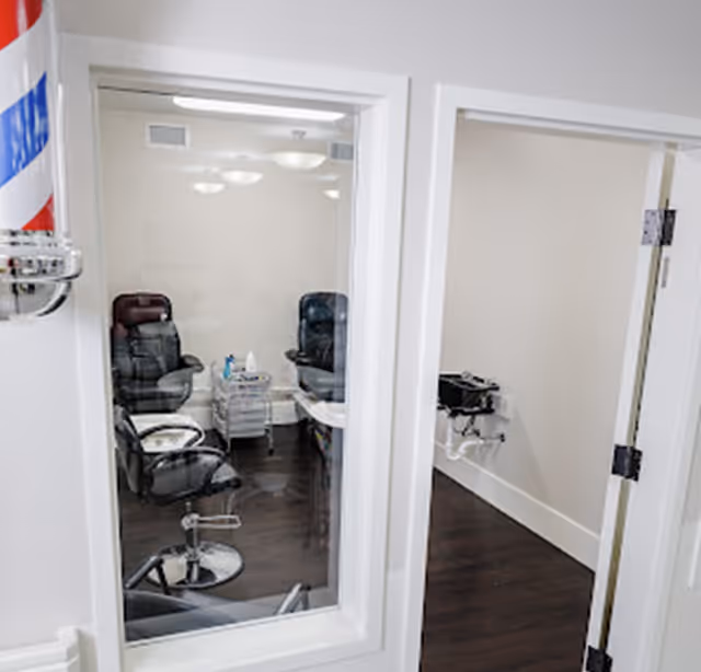 Interior view of a small barber or salon room with two black leather barber chairs, a small cart with supplies, and a white sink mounted on the wall. A traditional red, white, and blue barber pole is partially visible on the left side of the image.
