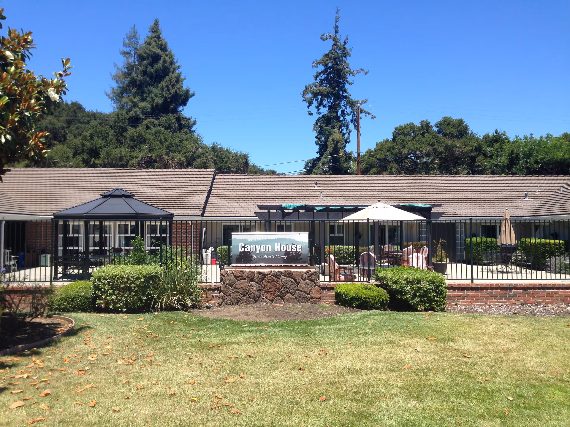 Outdoor view of a senior living facility named Canyon House with a fenced patio area featuring outdoor seating, umbrellas, and greenery. The building has a brown shingled roof and is surrounded by trees and bushes under a clear blue sky.