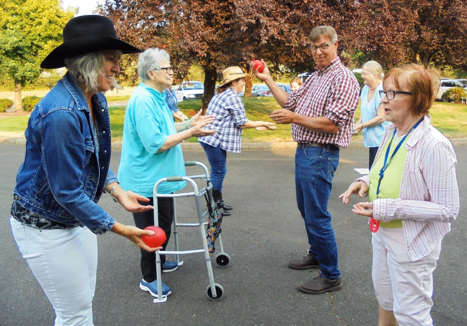 A group of elderly people and a man are playing a game with red balls outdoors in a parking lot area. Some participants are using walkers, and they are all casually dressed, enjoying the activity under trees with autumn-colored leaves.
