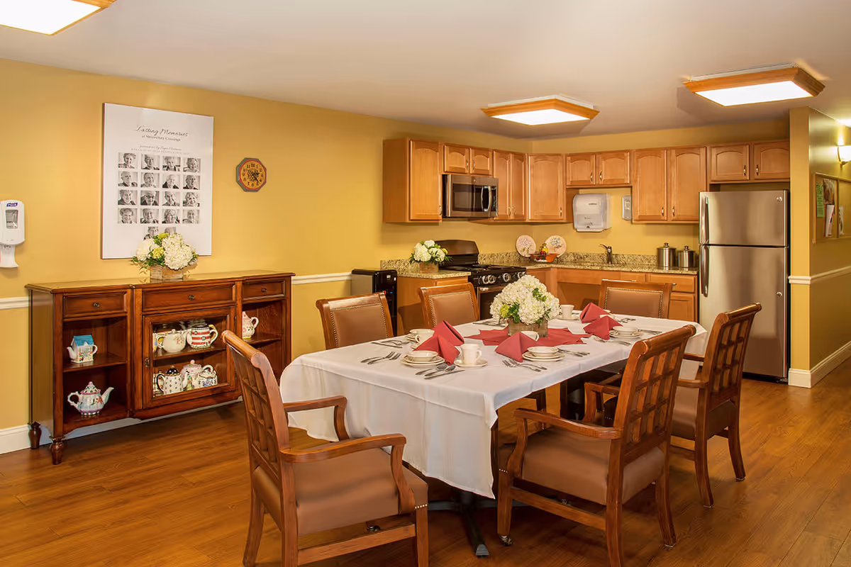 Dining room with a table set for a meal, wooden chairs and a sideboard, adjacent to an open kitchen with wooden cabinets and a stainless refrigerator.