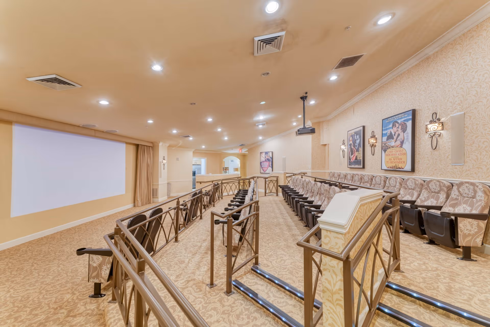 Interior view of a small theater room with rows of cushioned seats arranged on a sloped floor facing a large blank projection screen. The walls are decorated with framed movie posters and patterned wallpaper, and there are wall-mounted lights providing soft illumination. The room has a beige and brown color scheme with carpeted floors and metal railings along the aisles.