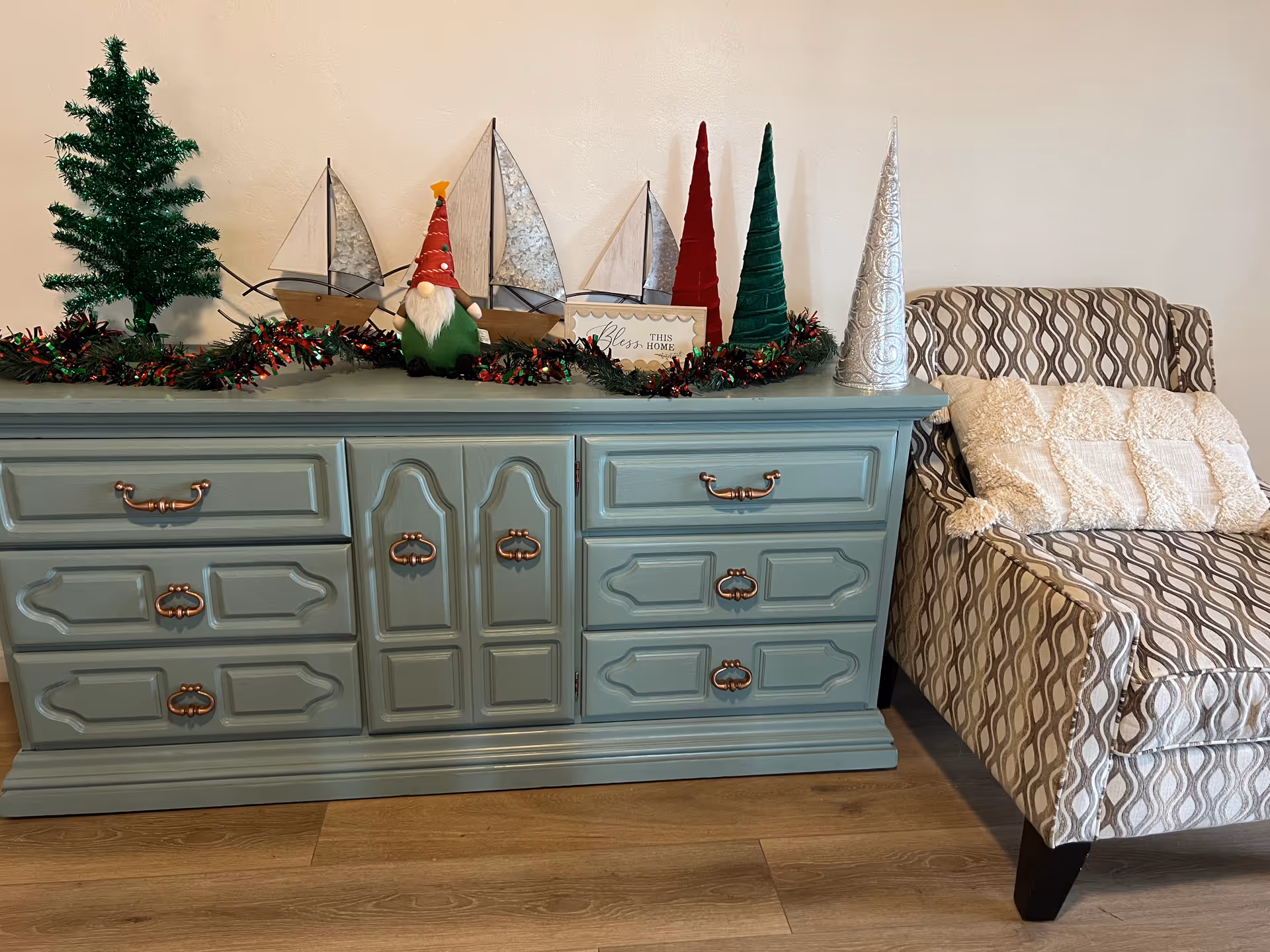 A light blue wooden dresser with decorative handles is topped with holiday decorations including a small green Christmas tree, three sailboat models, a gnome figurine wearing a red hat, a garland with red and green tinsel, a small framed sign that says 'Bless this home', and three cone-shaped Christmas trees in red, green, and silver. To the right of the dresser is a patterned armchair with a cream-colored textured pillow. The floor is wooden and the wall behind is plain white.