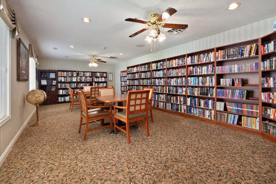 A well-lit library room with large wooden bookshelves filled with books along the walls. There are two wooden tables with chairs arranged around them in the center of the room. The room has patterned carpet flooring, ceiling fans with lights, and windows with blinds and valances on the left side. A globe stands near the window.