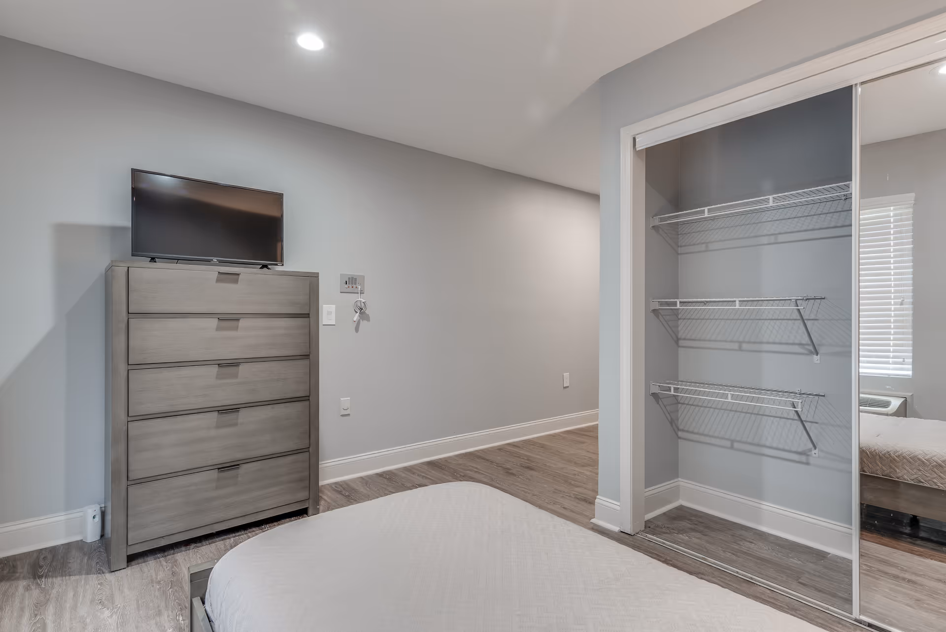 A modern bedroom with a tall gray dresser topped by a TV, an open closet with wire shelves, and part of a bed in a neutral gray interior.