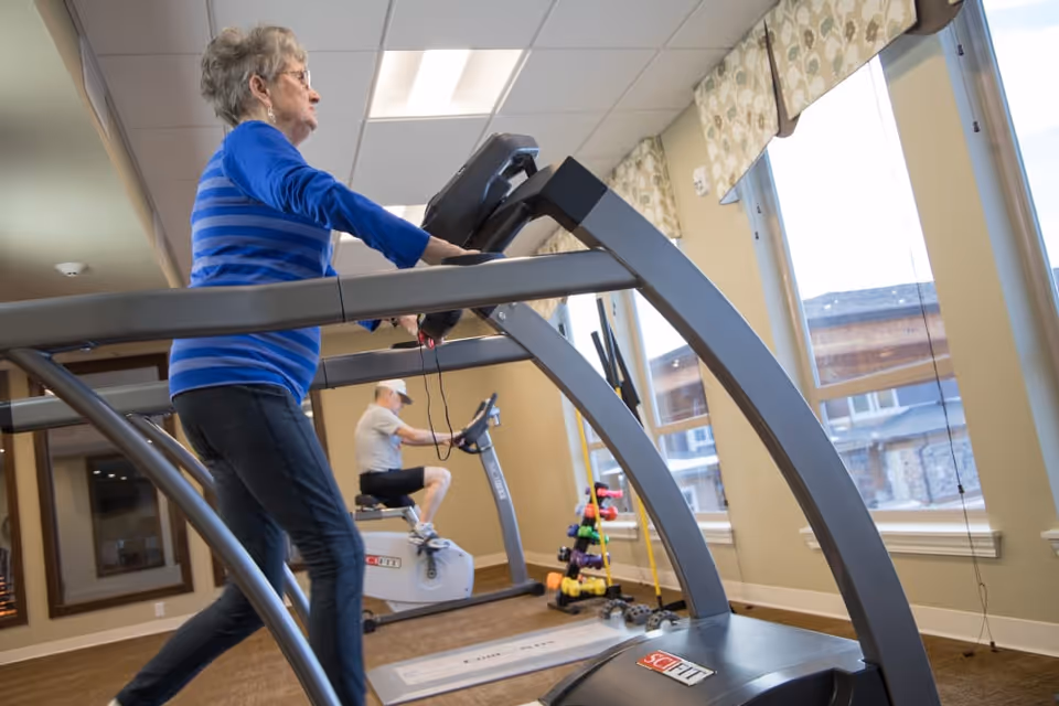 An elderly woman walking on a treadmill in a fitness room with large windows. In the background, an elderly man is using a stationary exercise bike. The room has light-colored walls, patterned window valances, and a rack of colorful dumbbells near the windows.