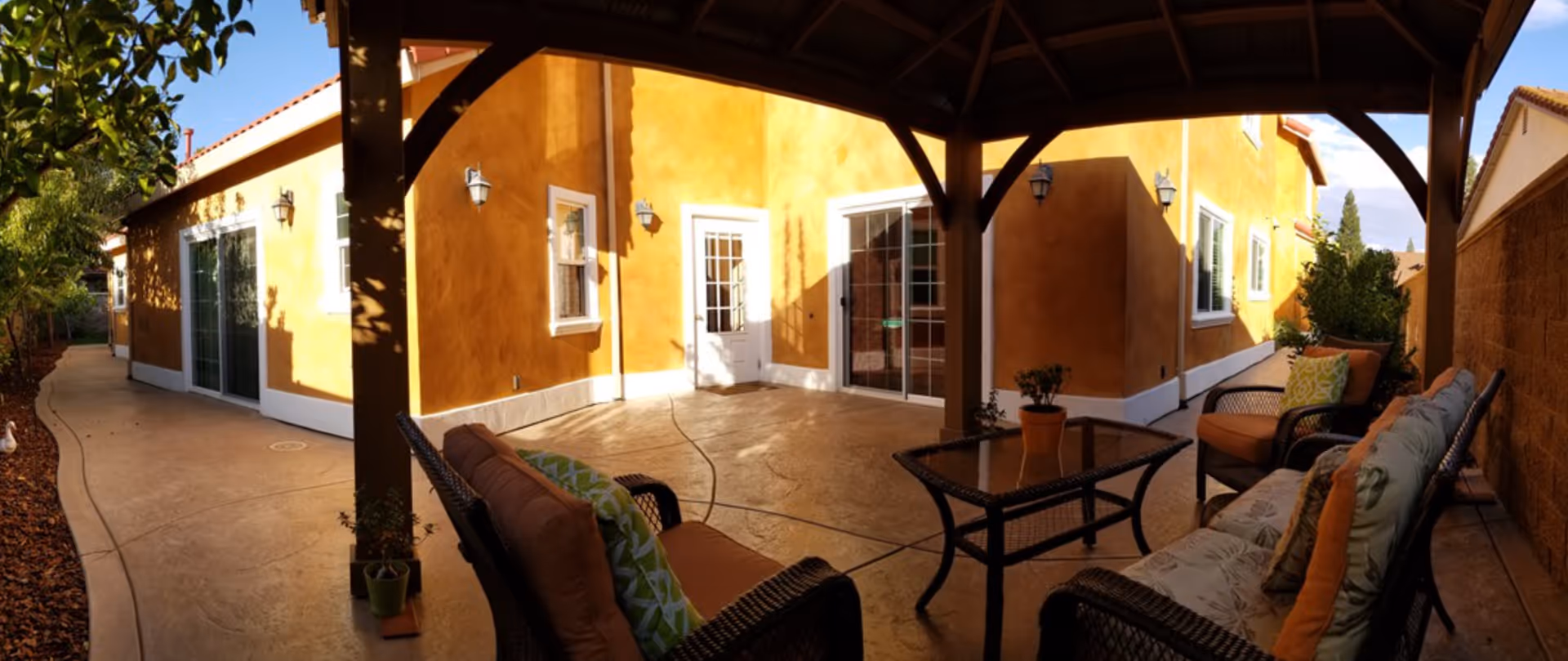 Covered patio with wicker seating and a glass-top table in front of a stucco house under a wooden pergola.