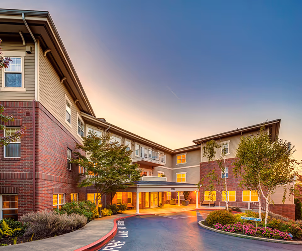 Exterior view of a multi-story senior living facility building at sunset with warm lights glowing from the windows. The building features a combination of brick and siding, with trees and landscaped bushes around the entrance driveway.