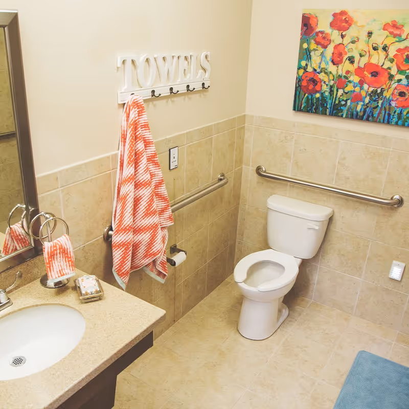 A bathroom with beige tiled walls and floor, featuring a white toilet with grab bars on both sides, a sink with a countertop, a mirror, and a towel rack with a red and white patterned towel hanging. There is a colorful painting of red flowers on the wall above the toilet.
