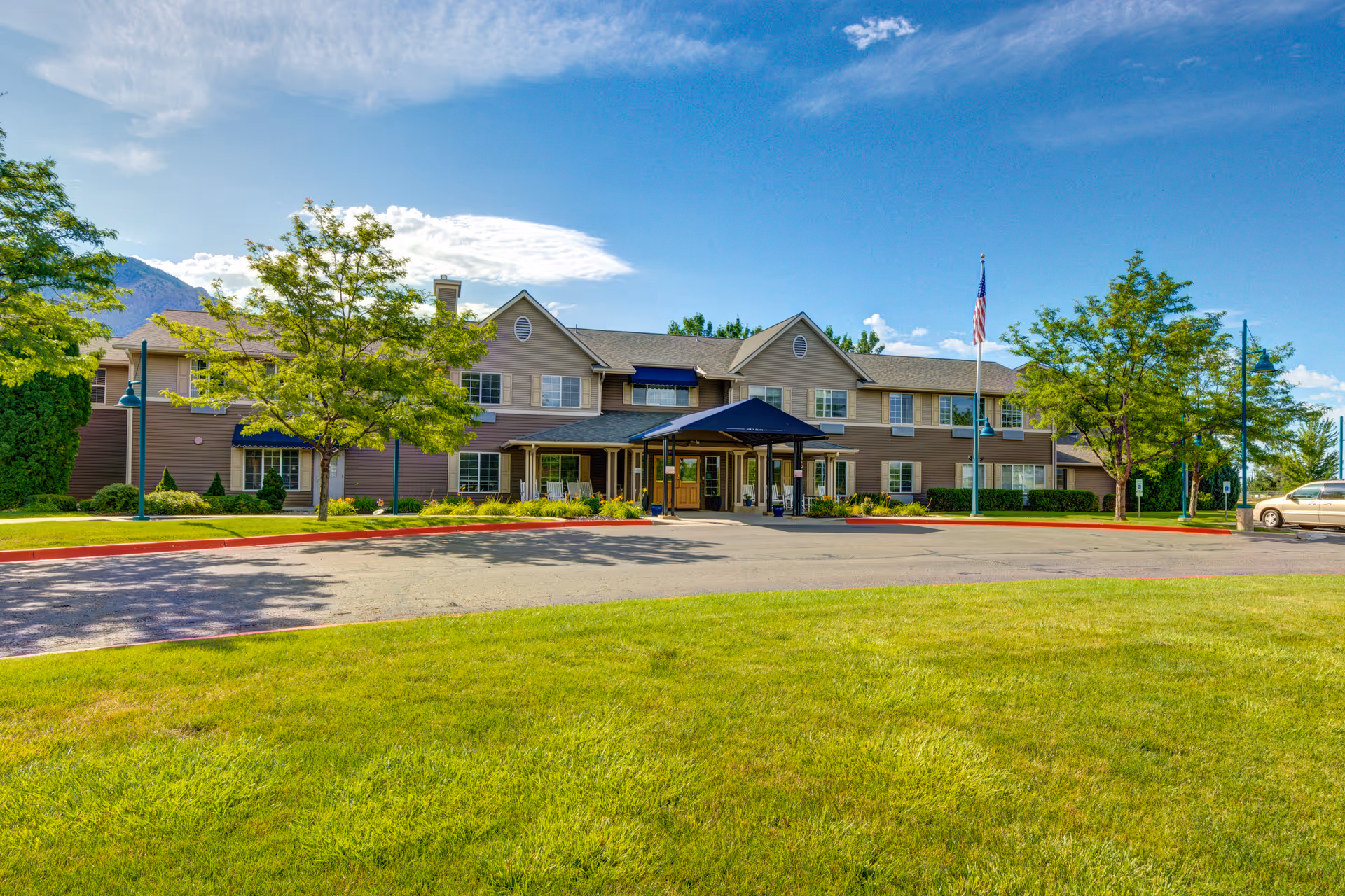 Exterior view of a two-story senior living facility building with a covered entrance, surrounded by green grass, trees, and a clear blue sky with some clouds.