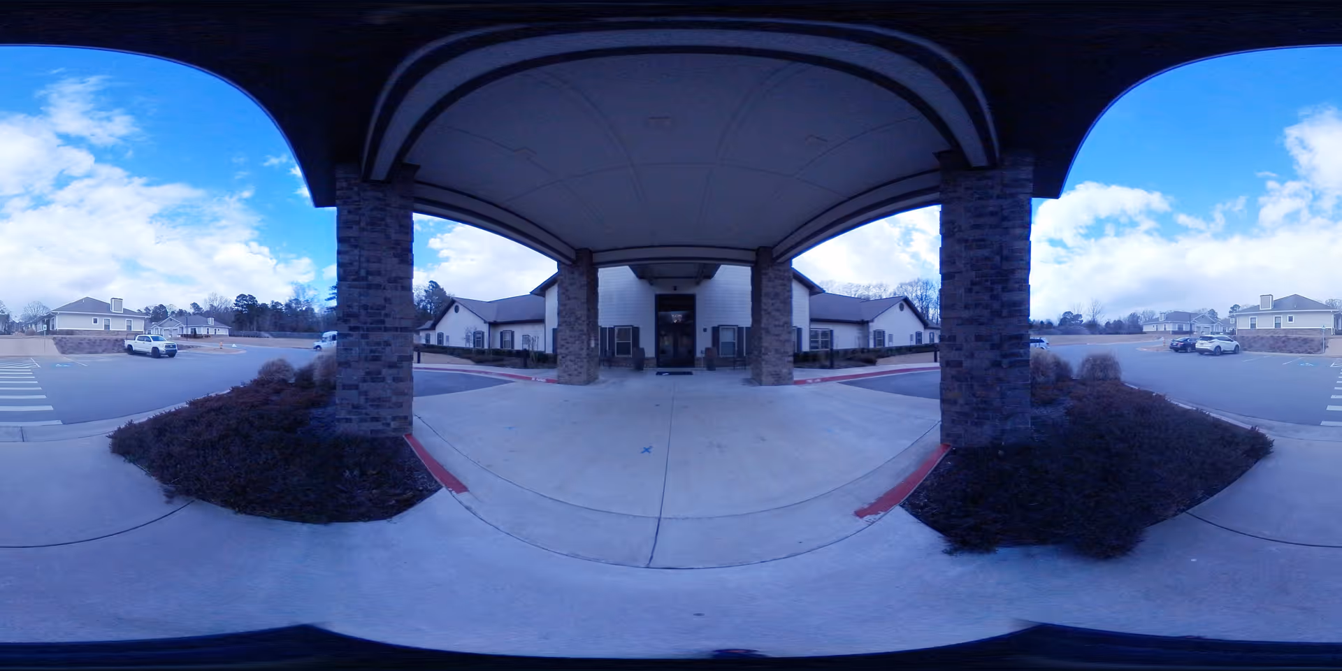 Covered entrance area of a senior living facility with stone pillars supporting a roof, leading to the main building entrance. The surrounding area includes parking spaces, landscaped bushes, and additional buildings under a partly cloudy sky.