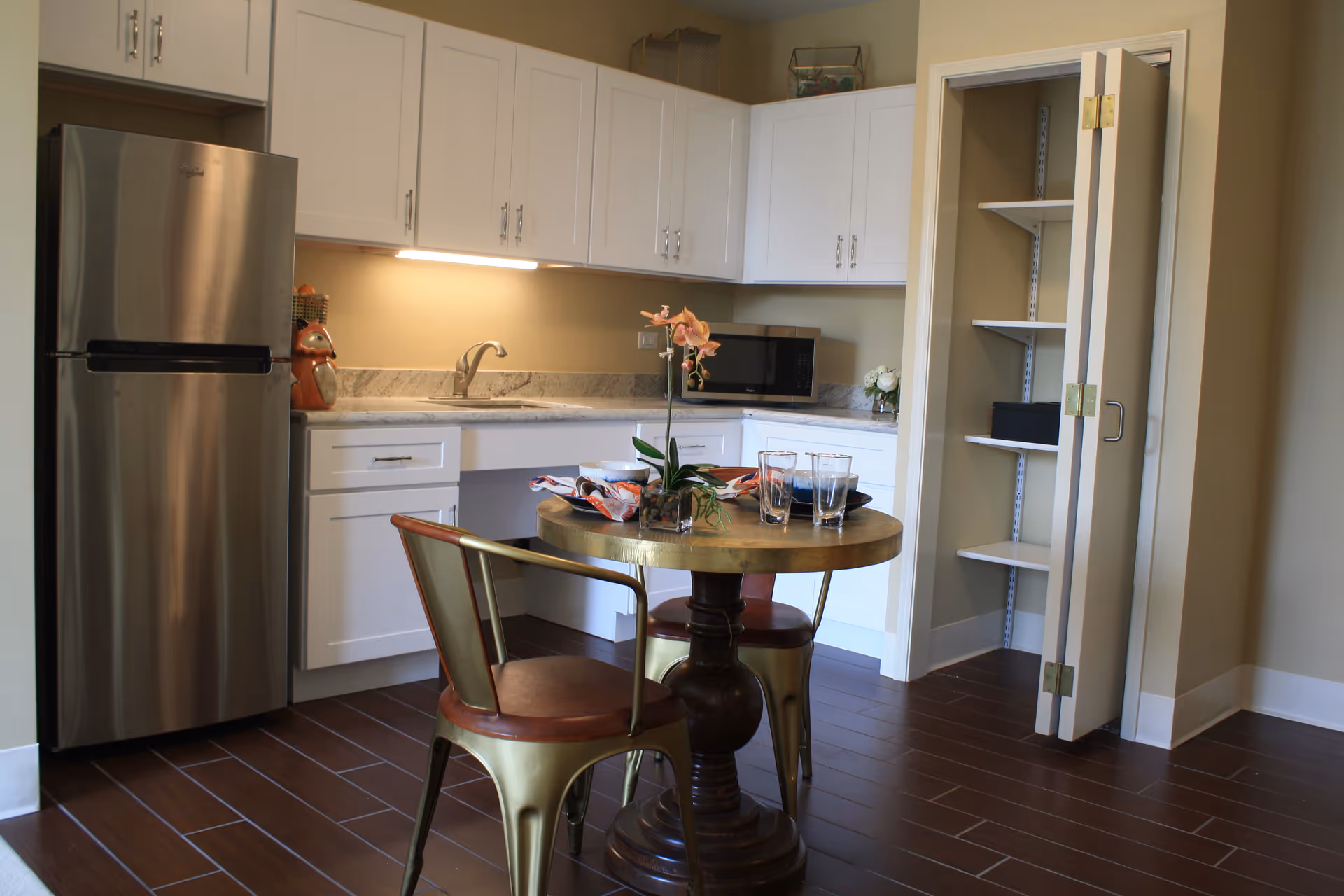 A small kitchen area with white cabinets, a stainless steel refrigerator, a microwave, and a sink. There is a round wooden table with two metal chairs, set with plates, glasses, and a small plant. An open pantry with shelves is visible to the right.