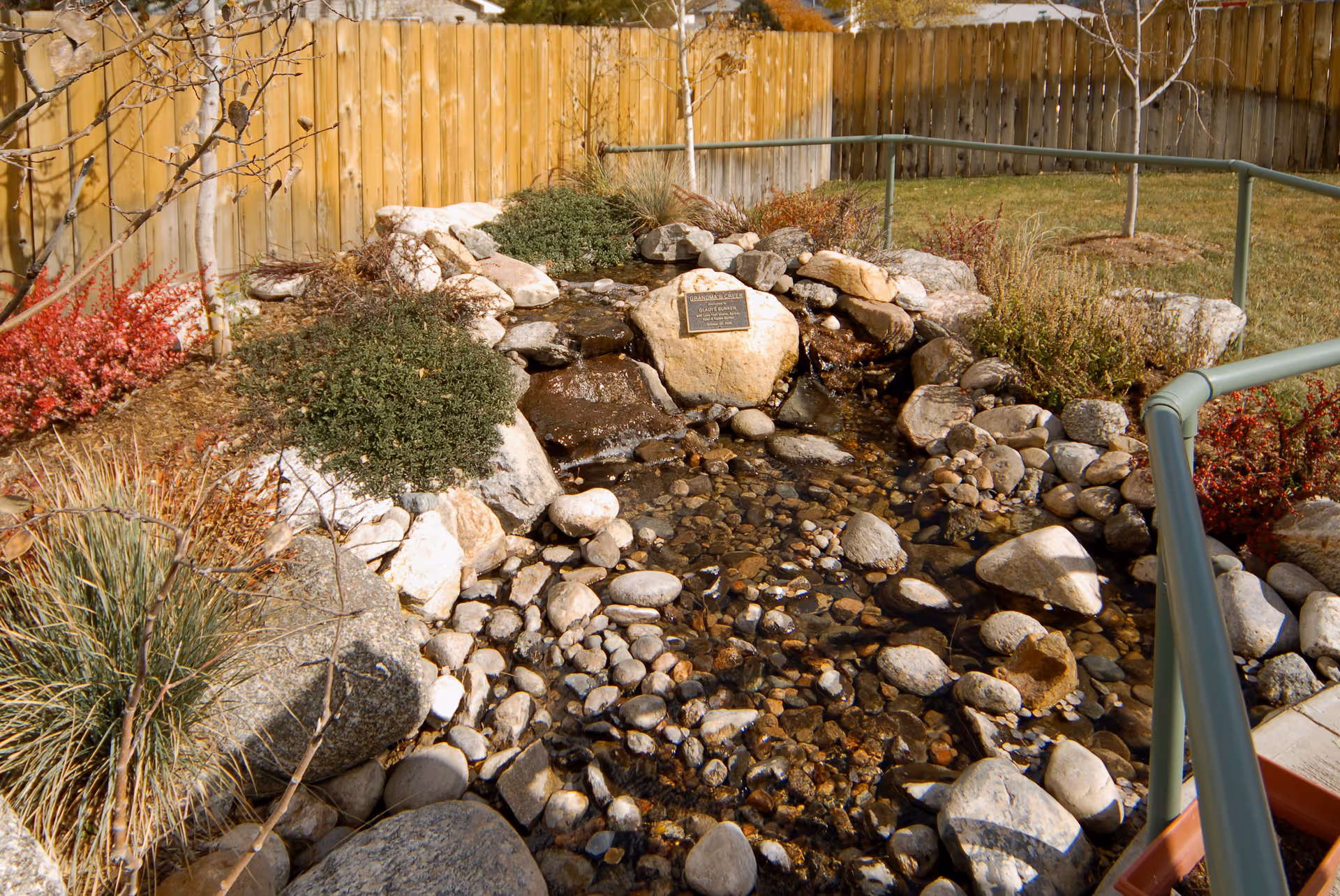 A small landscaped rock-lined pond and stream with shrubs, a commemorative plaque, a wooden fence and a green handrail.