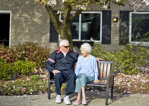 An elderly man and woman sitting together on a bench outside near a tree with blooming flowers, in front of a brick building with windows and shrubs.