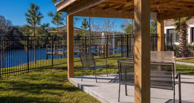Covered outdoor seating area with metal chairs and a bench under a wooden pergola beside a fenced lake and dock.