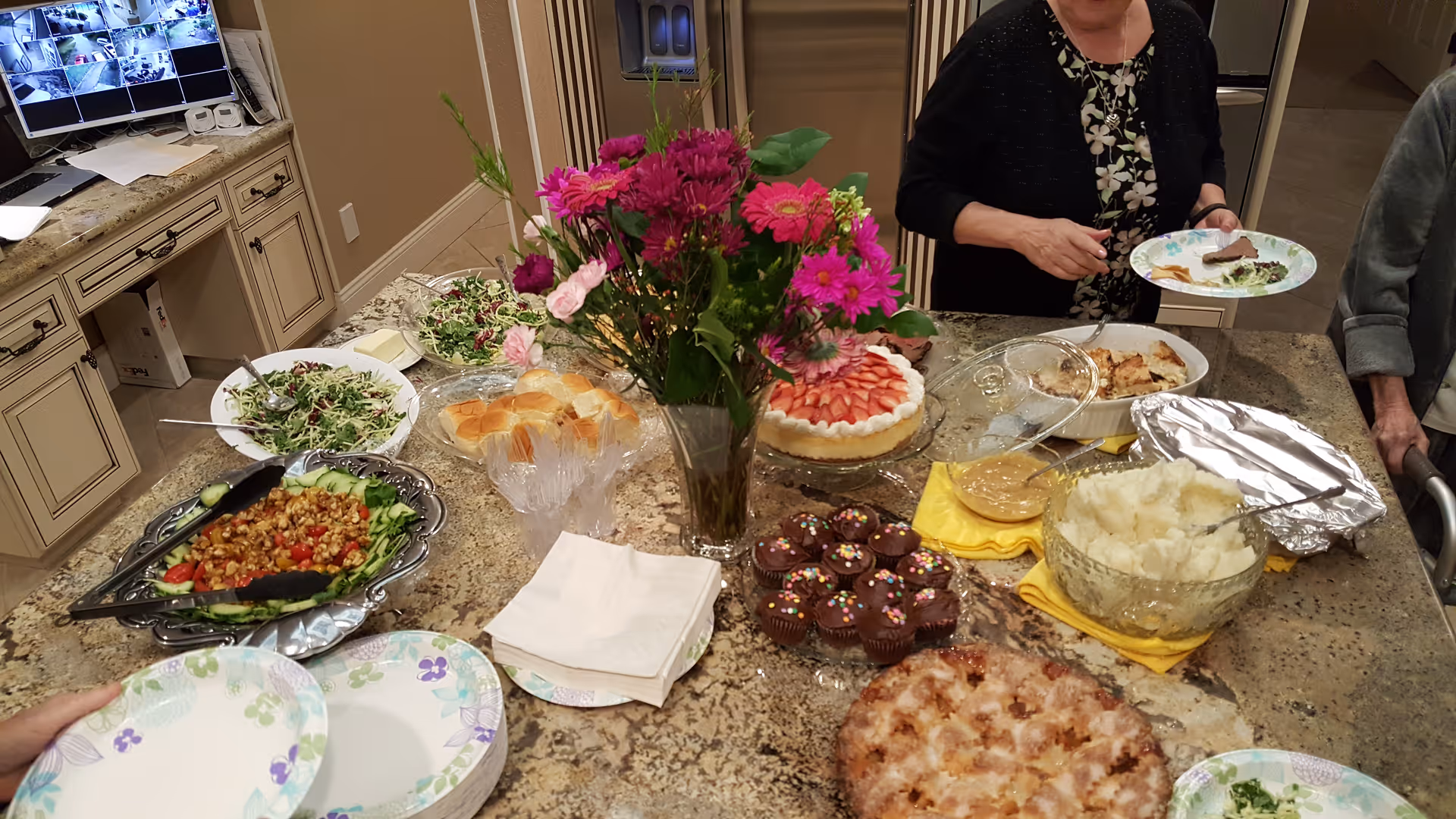 A kitchen countertop with a variety of food dishes including salad, bread rolls, mashed potatoes, cupcakes, a strawberry-topped dessert, and other prepared foods. Two elderly women are serving themselves from the food. A vase with pink and purple flowers is placed in the center of the countertop.