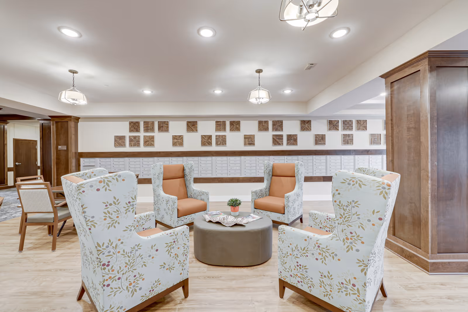 A bright and spacious seating area with four floral-patterned armchairs and two solid orange armchairs arranged around a round ottoman with a small plant and magazines on it. The background features a wall with multiple mailboxes and decorative wooden square panels above them. The room has wooden flooring, white walls, and recessed ceiling lights with two hanging light fixtures.