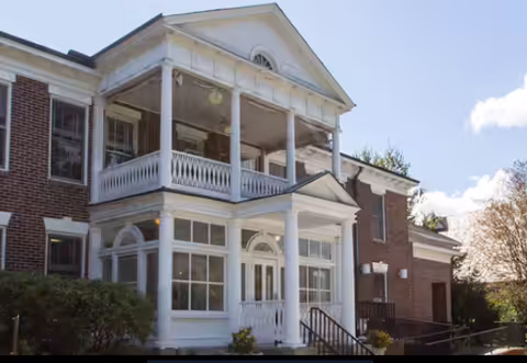 Exterior view of a two-story brick building with white columns and a covered porch area. The building has multiple windows and a small set of stairs leading to the entrance. There are bushes and trees around the building under a partly cloudy sky.