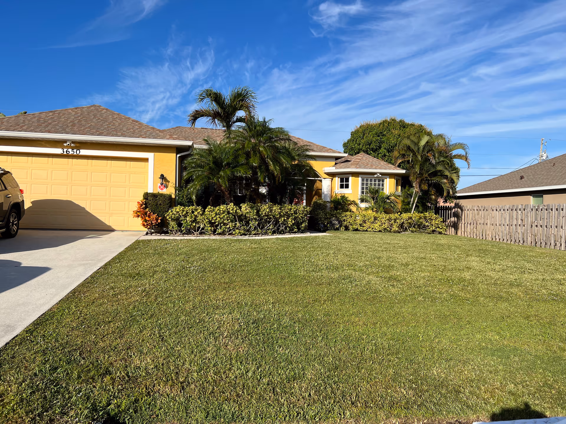 Front view of a yellow single-story house with a two-car garage, palm trees, and a green lawn under a blue sky.