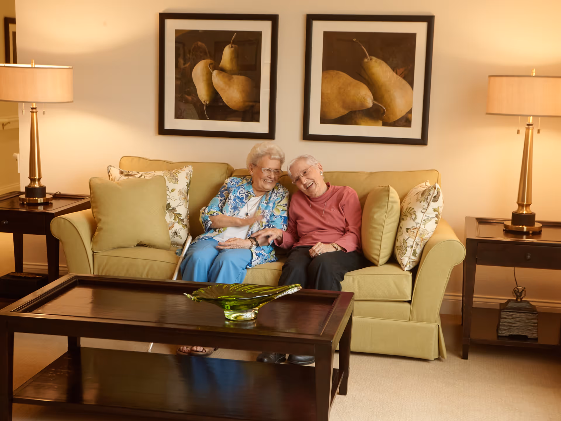 Two elderly women sitting closely together on a green sofa in a warmly lit living room, smiling and holding hands. The room features two framed pictures of pears on the wall behind them, two side tables with matching lamps, and a dark wooden coffee table with a green decorative glass piece.