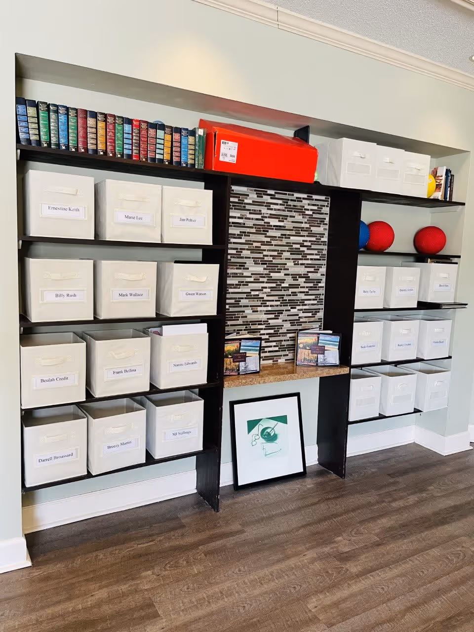 A shelving unit with multiple white storage bins labeled with names, a row of colorful books on the top left shelf, a large red box on the top middle shelf, and two red and one blue balls on the right shelves. The middle section has a mosaic tile backsplash with a small countertop holding framed pictures, and a framed artwork resting on the floor below.