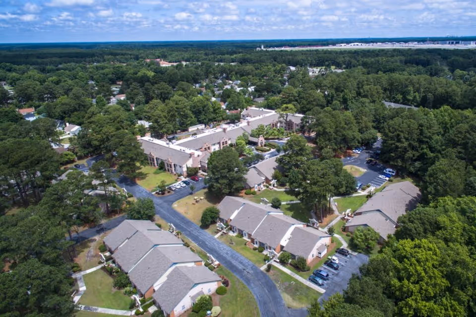 Aerial view of a residential senior living campus with multiple low-rise buildings, parking areas and surrounding trees.