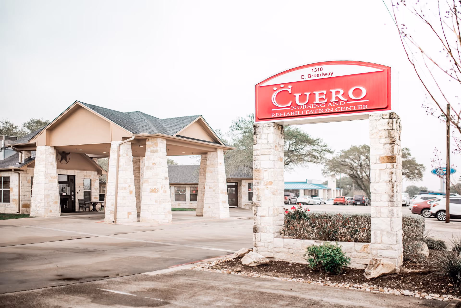 Exterior view of Cuero Nursing and Rehabilitation Center showing the entrance with stone pillars and a large red sign displaying the facility's name and address at 1310 E. Broadway. Several cars are parked in the background.
