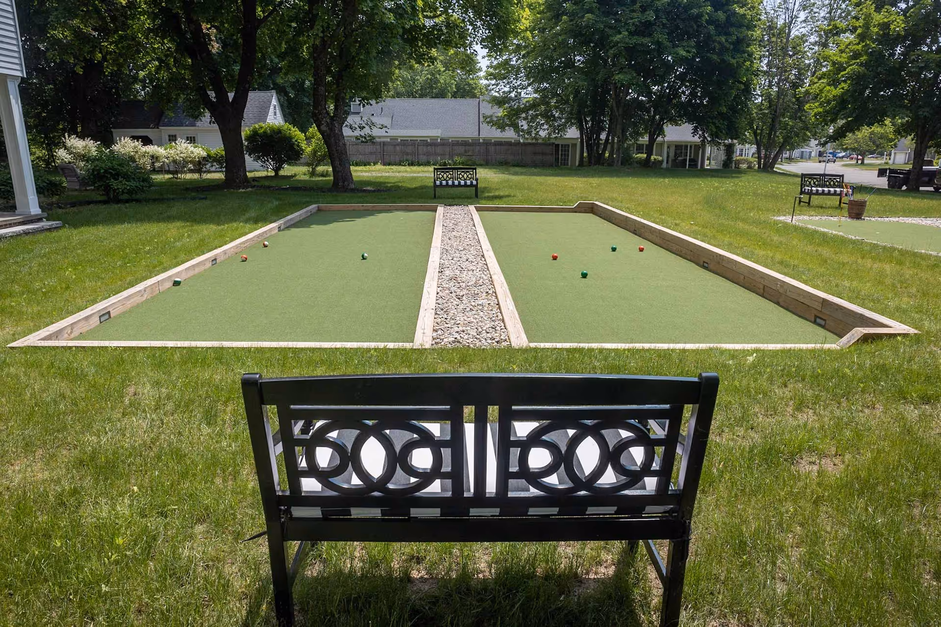 Outdoor bocce ball courts with green playing surfaces and wooden borders, surrounded by grass and trees. There are benches placed near the courts for seating, and houses are visible in the background.