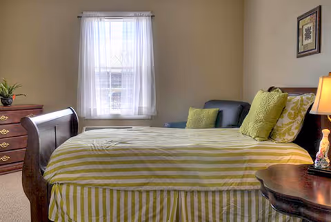 A bedroom with a wooden bed frame and a bed made with green and white striped bedding. There are two green pillows and one patterned pillow on the bed. Next to the bed is a wooden nightstand with a lamp and a decorative item. A window with sheer white curtains lets in natural light. A wooden dresser with a plant on top is visible on the left side of the room. The walls are painted beige and there is a framed picture hanging on the wall.