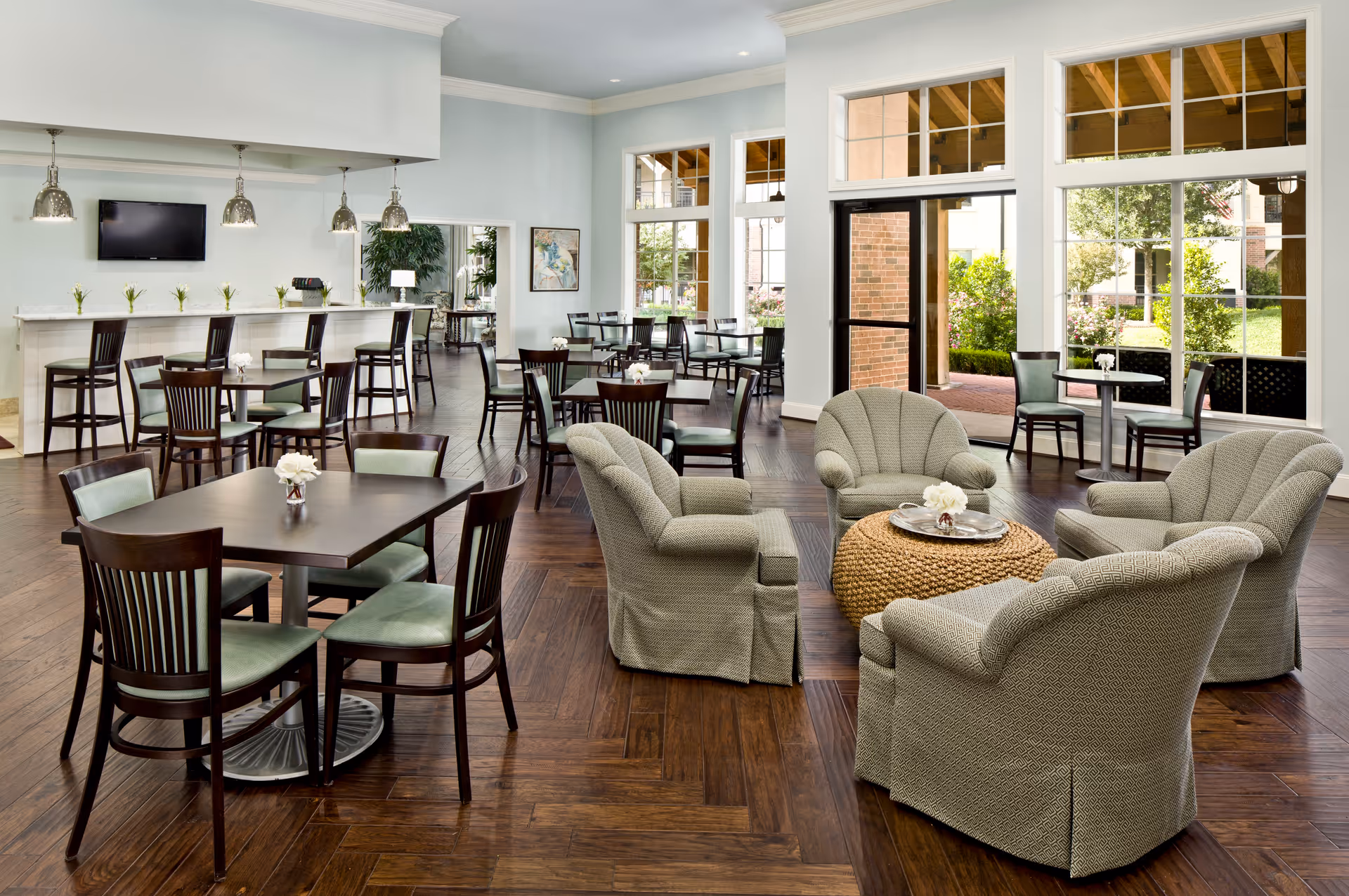 A bright and spacious common area with multiple seating options including a group of four upholstered armchairs arranged around a woven round coffee table, and several dark wood dining tables with matching chairs. Large windows and glass doors provide a view of a garden outside, allowing natural light to fill the room. A bar counter with high chairs and pendant lights is visible in the background, along with a wall-mounted TV and decorative plants.
