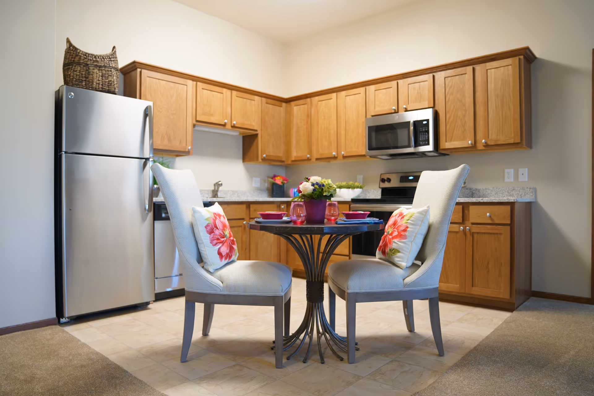 A cozy kitchen area with wooden cabinets, a stainless steel refrigerator, dishwasher, stove, and microwave. In the center, there is a small round dining table set for two with floral cushions on the chairs and a flower arrangement on the table.