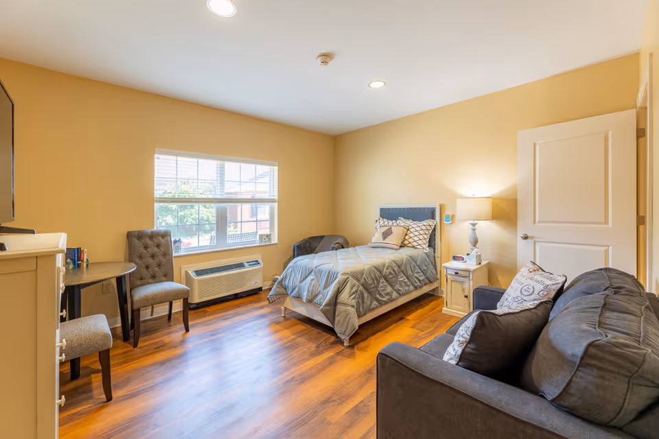 A cozy bedroom with a single bed covered in a gray quilt and decorative pillows. The room features a small white nightstand with a lamp, a gray armchair, a gray sofa with pillows, a round table with two chairs, a window with blinds, and wooden flooring.