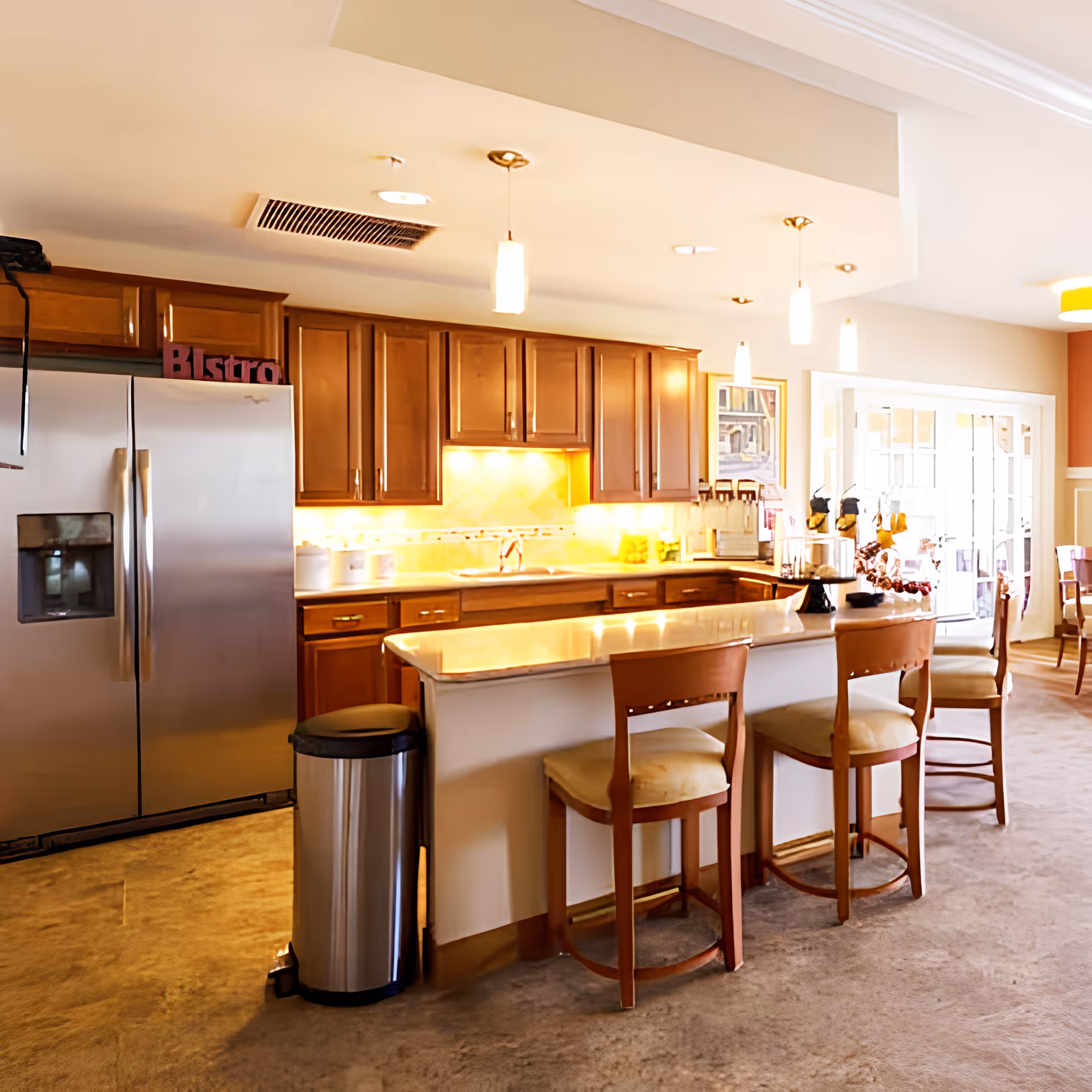 A bright and spacious kitchen area with wooden cabinets, a large stainless steel refrigerator, a kitchen island with a marble countertop, and three wooden chairs with cushioned seats. The kitchen is well-lit with pendant lights hanging from the ceiling and under-cabinet lighting. There is a trash can next to the island and a dining area visible in the background with large windows letting in natural light.
