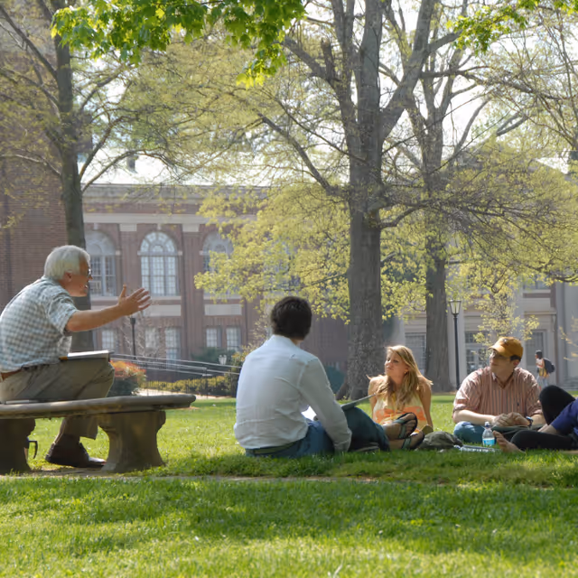 A small group of people sitting and talking on a grassy lawn under trees in front of a large brick building.